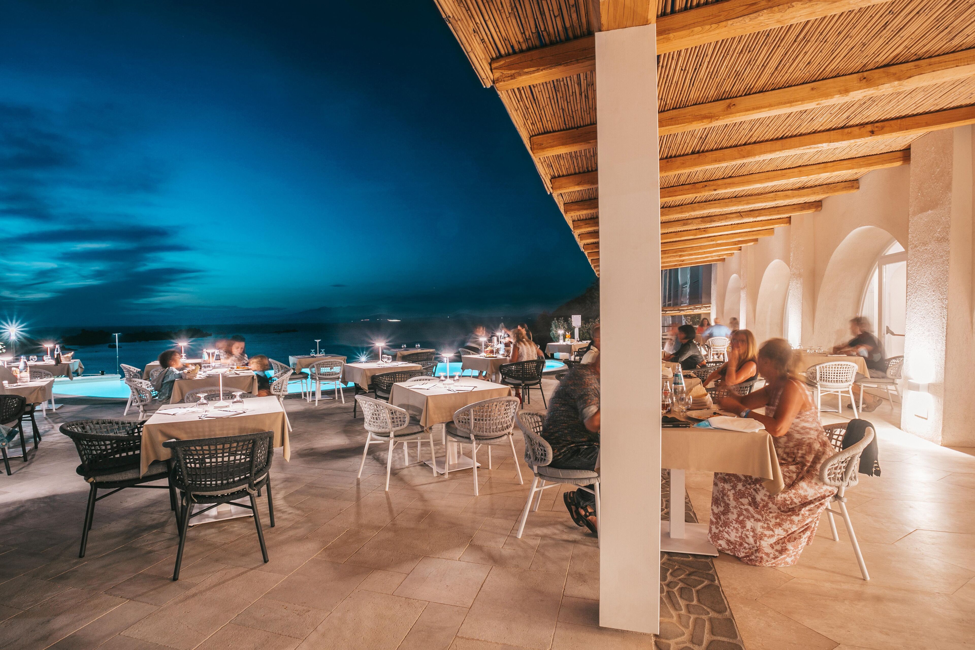 An outdoor dining area with tables and chairs, a wooden ceiling, and a view of the sea.
