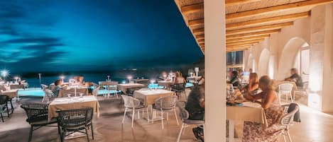 An outdoor dining area with tables and chairs, a wooden ceiling, and a view of the sea.