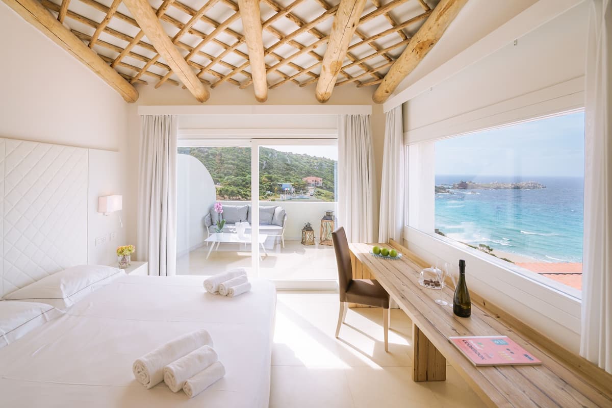 A hotel room with a bed, a wooden desk, a view of the sea, and a balcony with outdoor furniture.