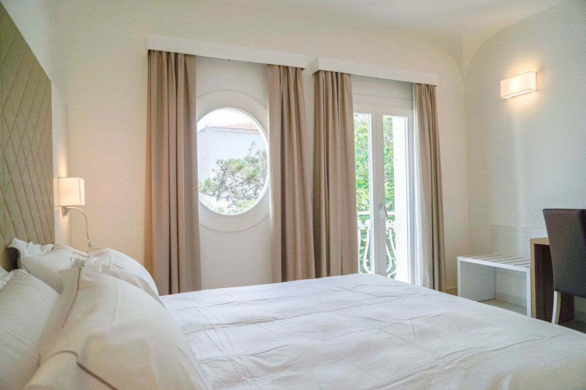 Una habitación de hotel con una cama grande, una ventana redonda, cortinas, un escritorio y una silla.