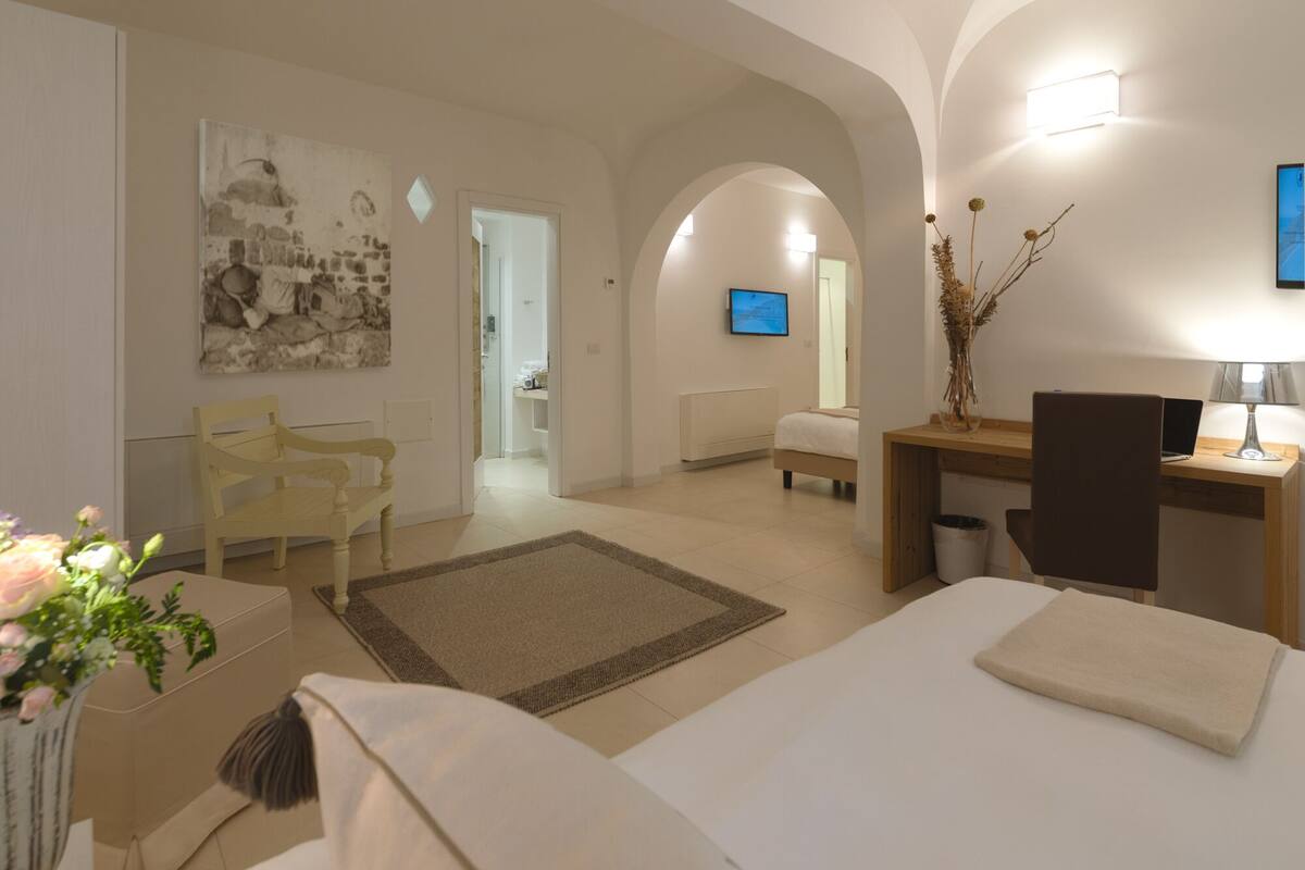 A hotel room with a bed, a desk, a chair, a small table, a vase with flowers, a picture on the wall, and a TV in the background.