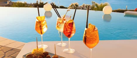 A table with four drinks and a plate of pastries by a poolside with a view of the sea.
