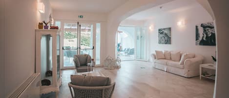 Amplia sala de estar con un sofá blanco, una silla de mimbre y un sillón blanco. Hay una puerta de vidrio que da a un espacio exterior.