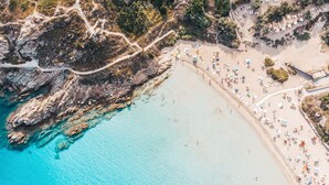 Vista aérea de una playa con aguas turquesas cristalinas, acantilados rocosos y una orilla arenosa con sombrillas y sillas de playa.