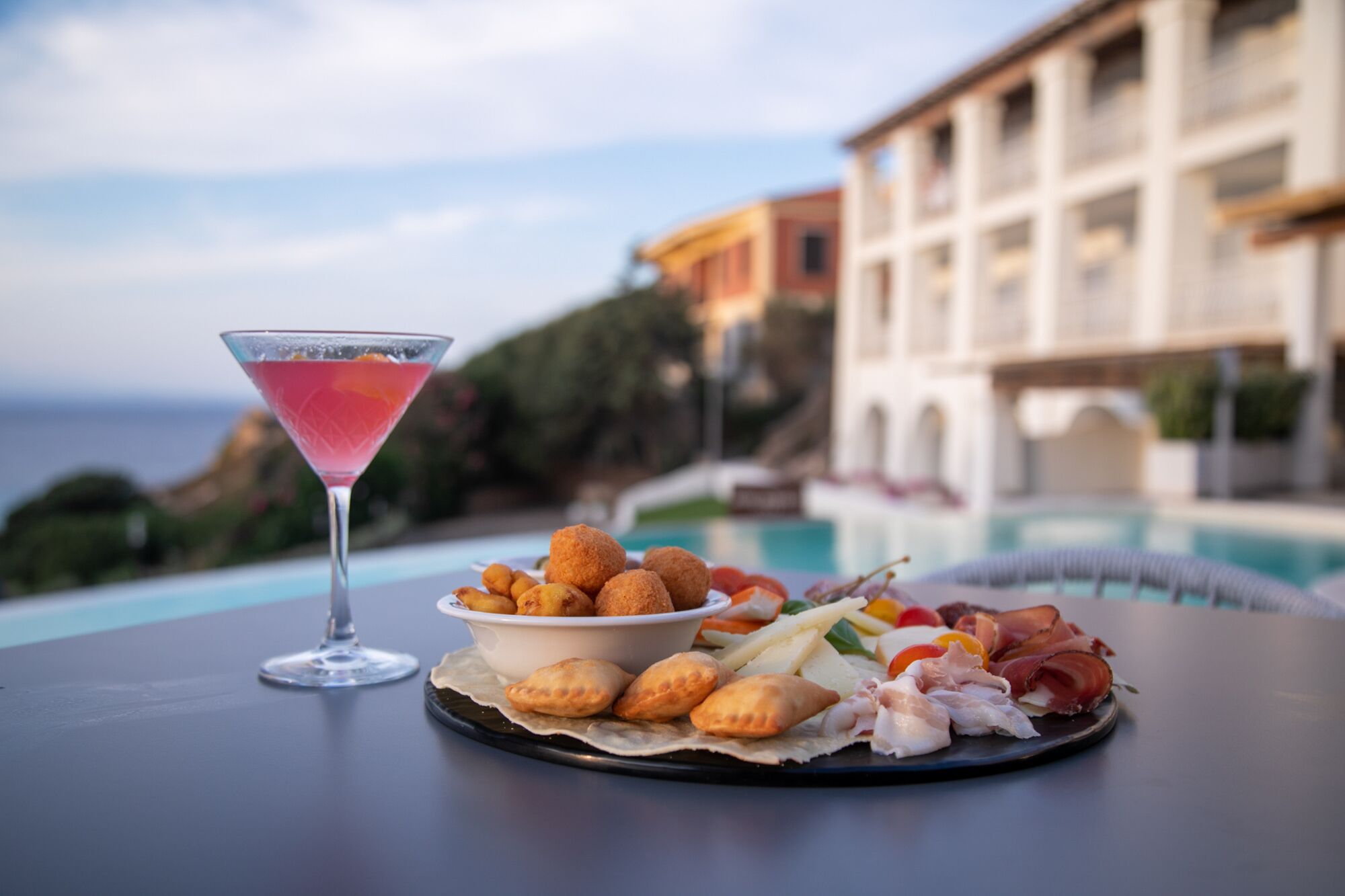A table with a martini glass, a bowl of appetizers, and a plate of food by a poolside with a building in the background.