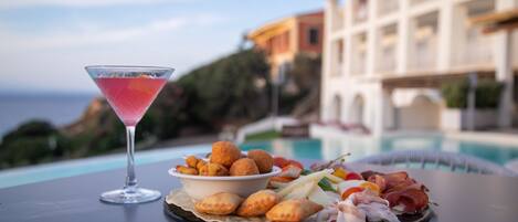 Una mesa con una copa de martini, un bol de aperitivos y un plato de comida junto a una piscina, con un edificio al fondo.