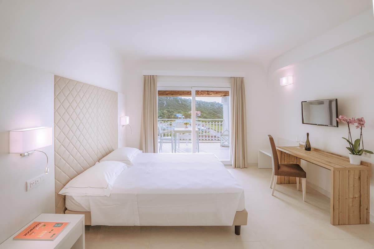 A hotel room with a bed, a desk, a chair, a TV, and a balcony with a view of the mountains.