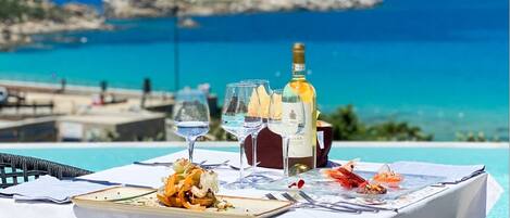 Une table dressée pour un repas, avec des plats de fruits de mer, des verres à vin et une bouteille de vin, donnant sur une mer bleue et limpide et une côte rocheuse.
