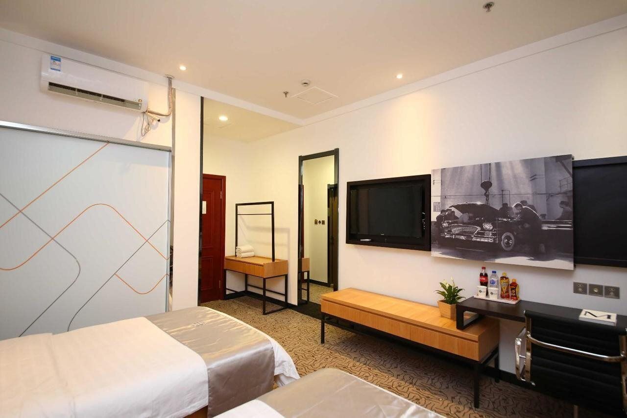 A hotel room with two beds, a TV, a mirror, a bench, and a wall-mounted TV.