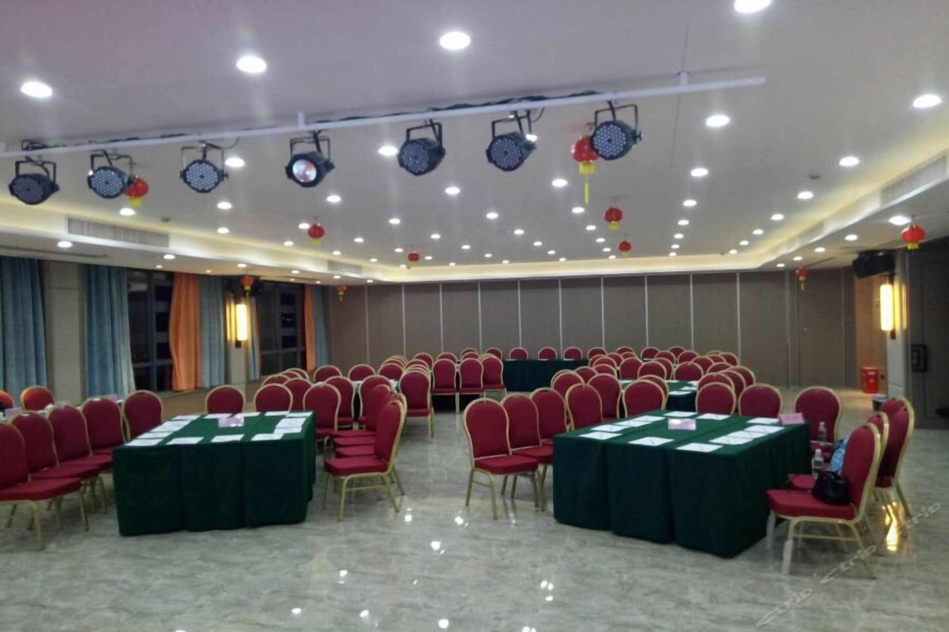 Un salón de conferencias con filas de sillas rojas y mesas verdes, equipado con luces en el techo y sistemas de sonido.