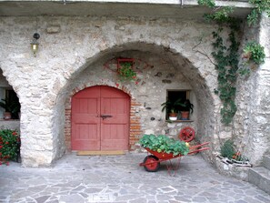 A stone building with a red door, a wheelbarrow with plants, and potted plants on the wall.