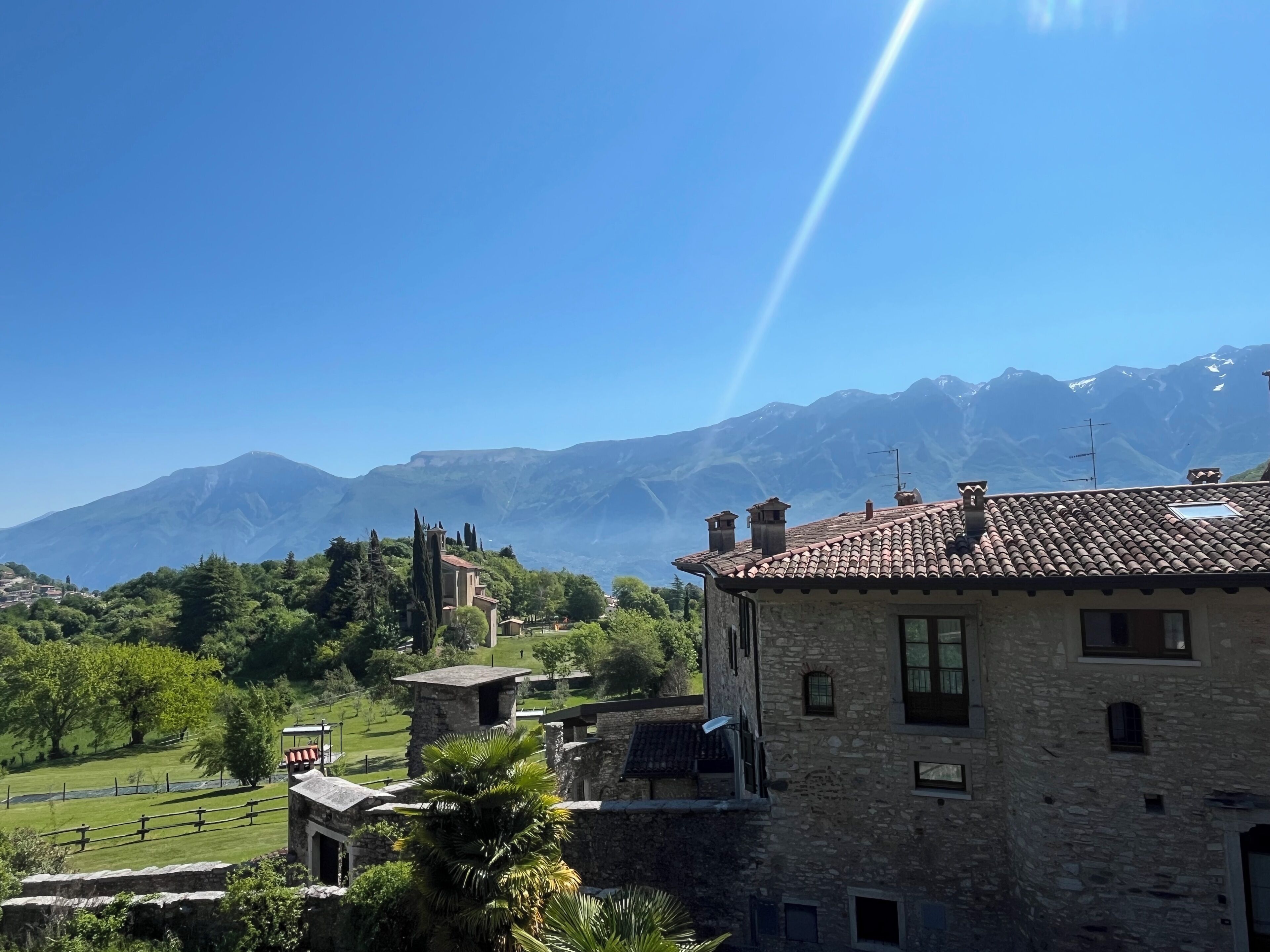 A stone building with a tiled roof, surrounded by greenery and mountains in the background.