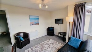 A living room with a sofa, a chair, a small table, a TV, and a painting of a beach. - Beachside Home - Sleeps 5, Garden, Parking & Pets (Skegness)