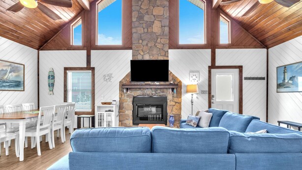 Ein großzügiger Wohnraum mit Kamin aus Stein, einem großen, blauen Sofa, Holzbalkendecke und einem Essbereich mit weißen Stühlen und einem Holztisch.