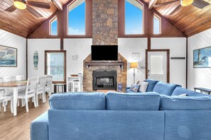 Een ruime woonkamer met een stenen openhaard, een grote blauwe bank, houten balken in het plafond en een eethoek met witte stoelen en een houten tafel.