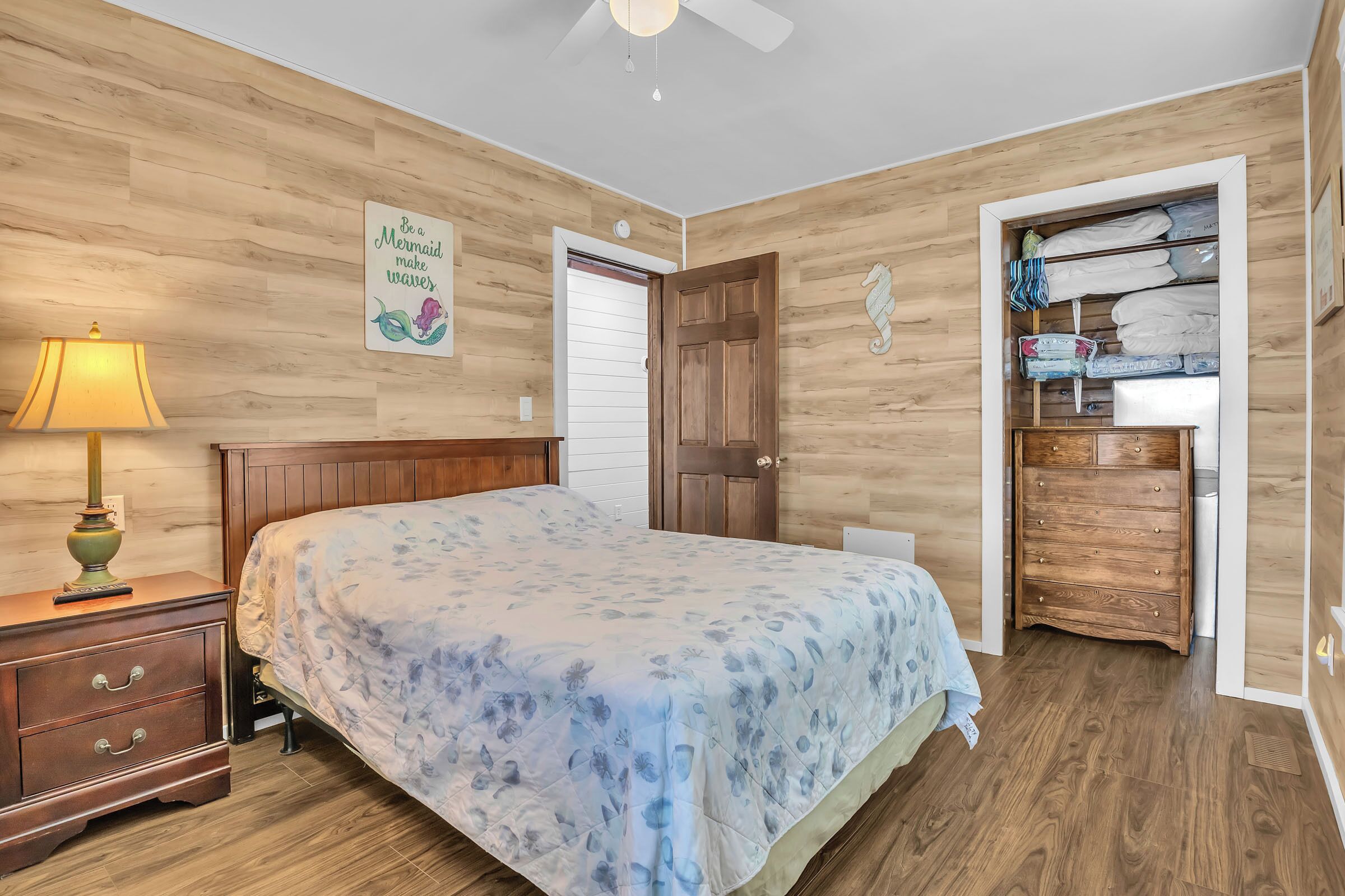 A bedroom with a wooden floor, a bed with a floral bedspread, a nightstand with a lamp, a wooden dresser, and a closet with folded clothes.