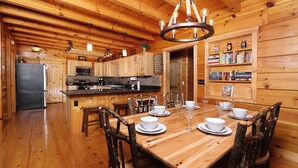 Interior de uma cabana de madeira com uma mesa de jantar posta para seis pessoas, uma cozinha com armários de madeira e uma estante repleta de livros.