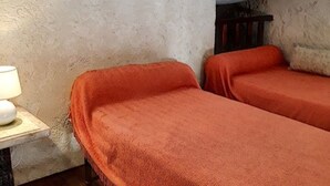Une petite pièce rustique avec deux lits simples recouverts de draps orange, une petite table avec une lampe et un mur à la texture particulière.