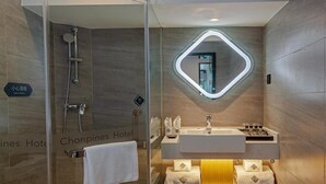 Un baño de hotel moderno con ducha envuelta en vidrio, un espejo grande con luz geométrica e un tocador con varios lavamanos y toallas.