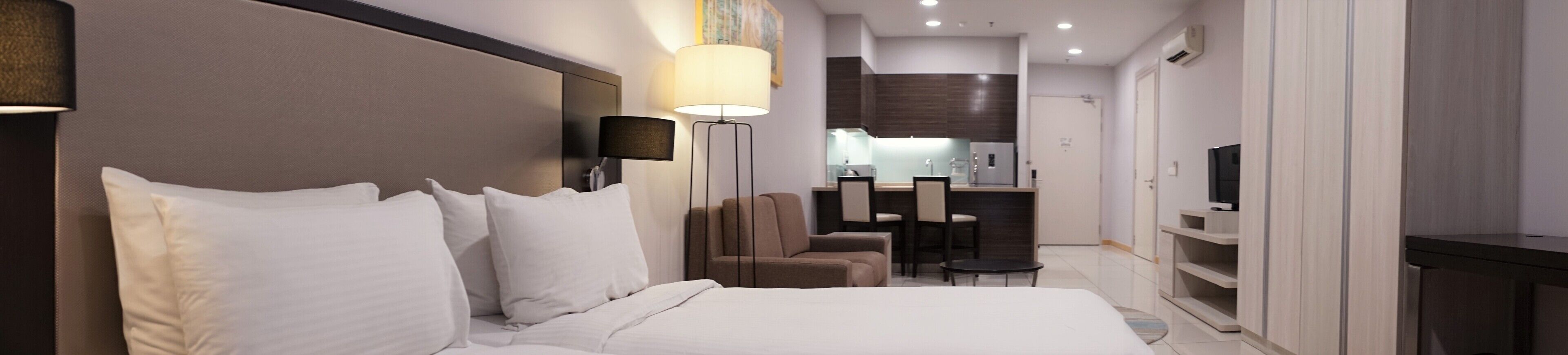A modern hotel room with a large bed, a desk, a chair, a TV, and a kitchenette.