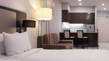 A modern hotel room with a large bed, a desk, a chair, a TV, and a kitchenette.