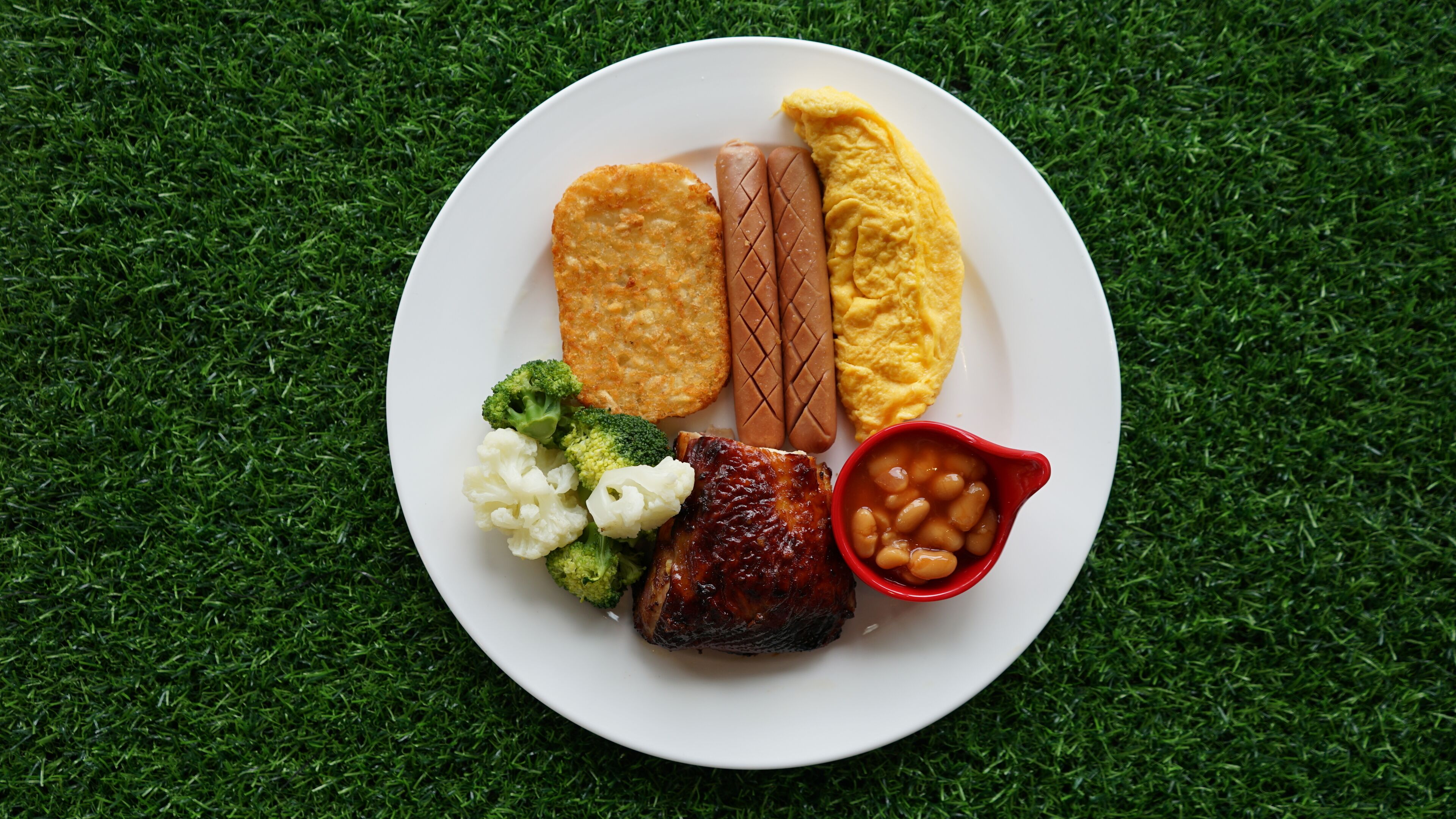 Un plato de comida con varios elementos, incluyendo una salchicha, un trozo de pollo frito, una patata al horno, una tostada y un tazón pequeño de frijoles.