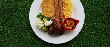 Un plato de comida con varios elementos, incluyendo una salchicha, un trozo de pollo frito, una patata al horno, una tostada y un tazón pequeño de frijoles.