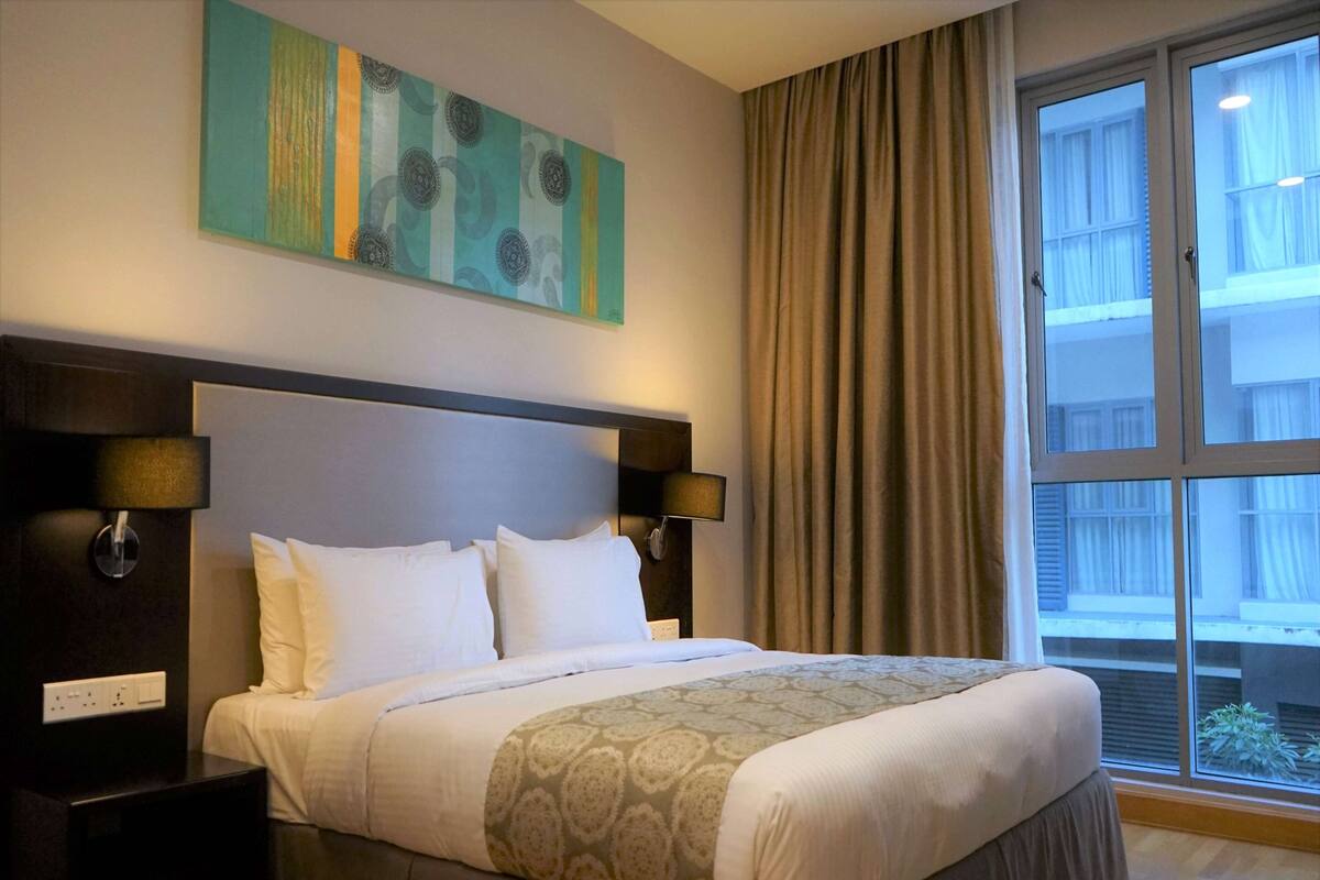 Una habitación de hotel con una cama grande, un cabecero con lámparas fijadas a la pared, una ventana con cortinas y un cuadro en la pared.