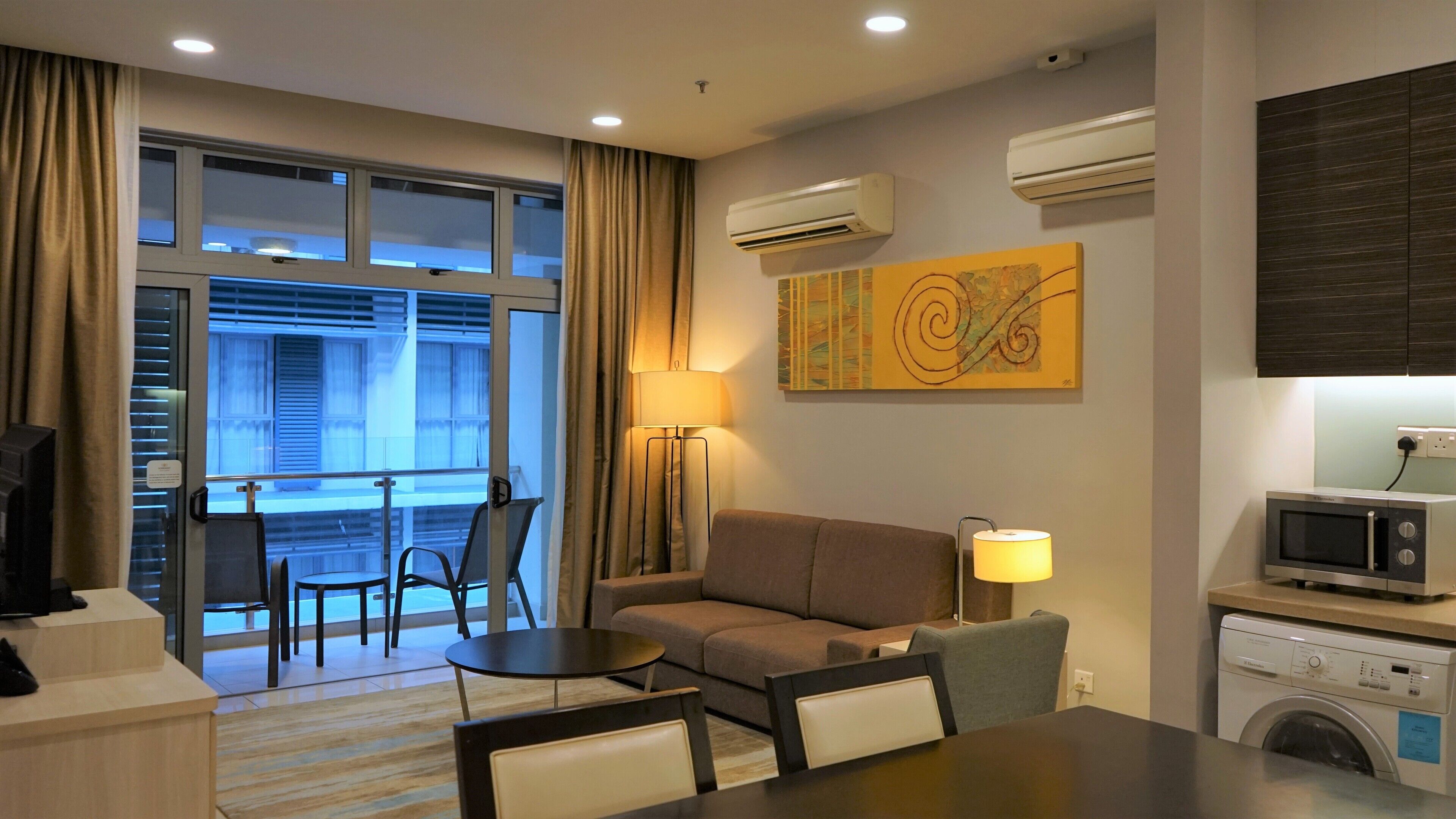 A hotel room with a sofa, a dining table, chairs, a TV, a washing machine, and an air conditioning unit.