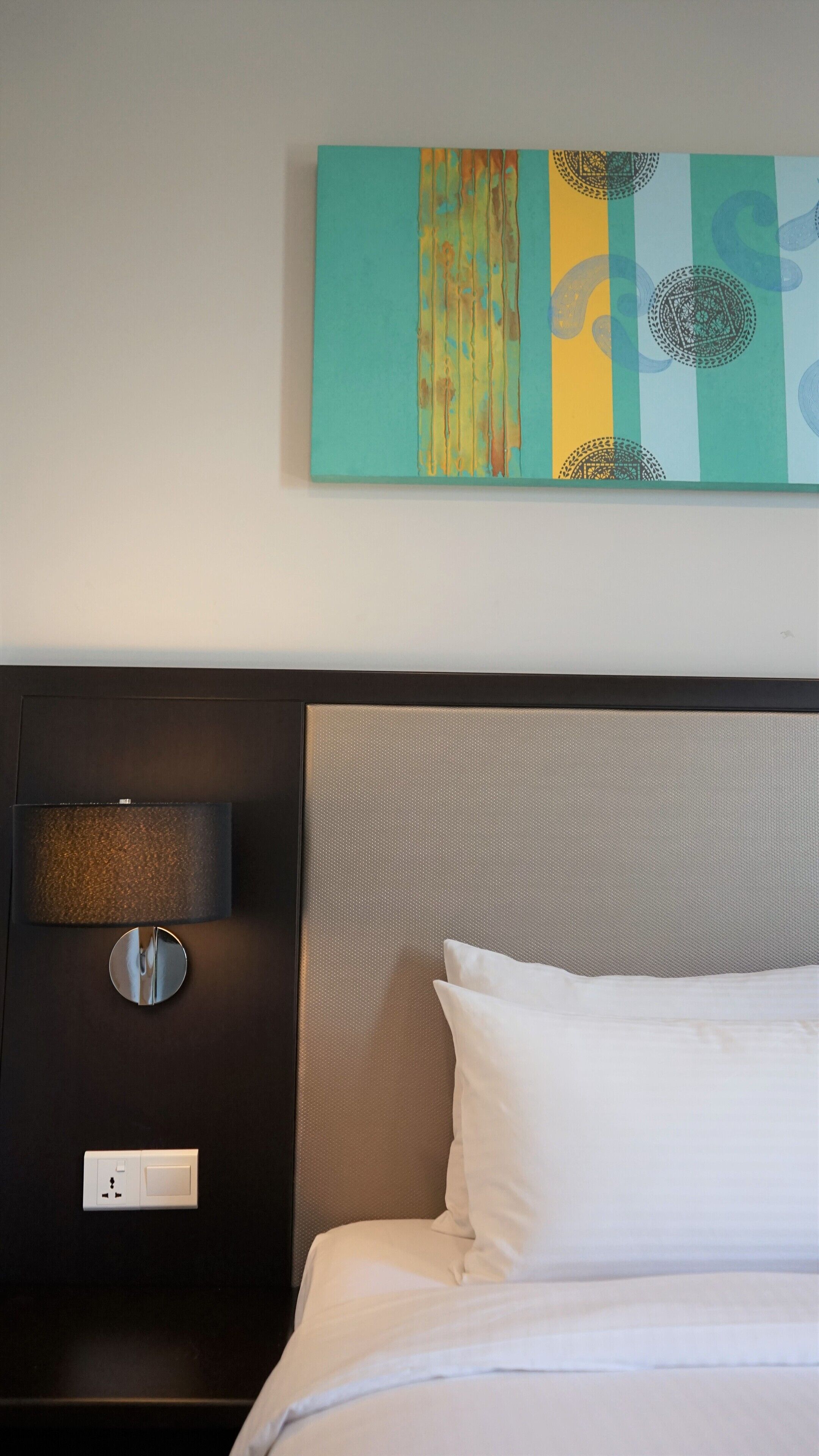 A hotel room with a bed, a wall-mounted lamp, and a painting above the bed.