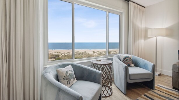 Una habitación con un gran ventanal con vista a la playa, dos sillones, una mesita redonda y una lámpara.