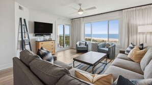 Amplia sala de estar con un televisor de pantalla plana grande, un ventilador de techo y vista a la playa a través de ventanas amplias.