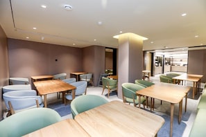 A modern interior with wooden tables and chairs, a kitchen area visible through an open doorway, and recessed ceiling lighting. - Ji Hotel (Chongqing)