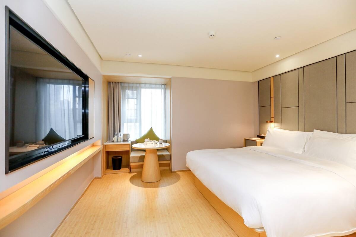 A modern hotel room with a large bed, a flat-screen TV mounted on the wall, a small round table with a green cushion, and a window with curtains.