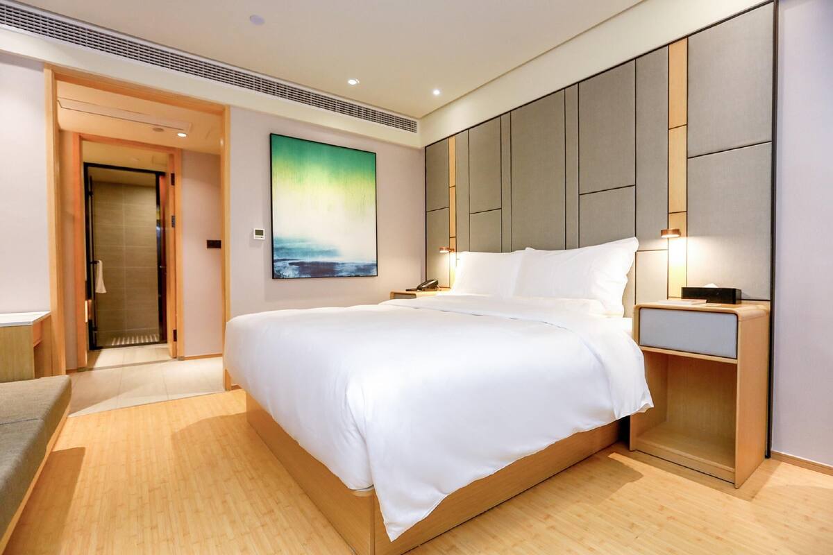 A modern hotel room with a large bed, wooden flooring, a painting on the wall, and a nightstand with a lamp.