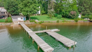 Un muelle de madera se extiende sobre un lago tranquilo, rodeado de abundante vegetación y un pequeño edificio blanco.