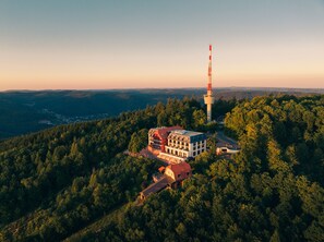 Un hotel con techo rojo y varios pisos, rodeado de un bosque denso y una alta torre de comunicaciones.