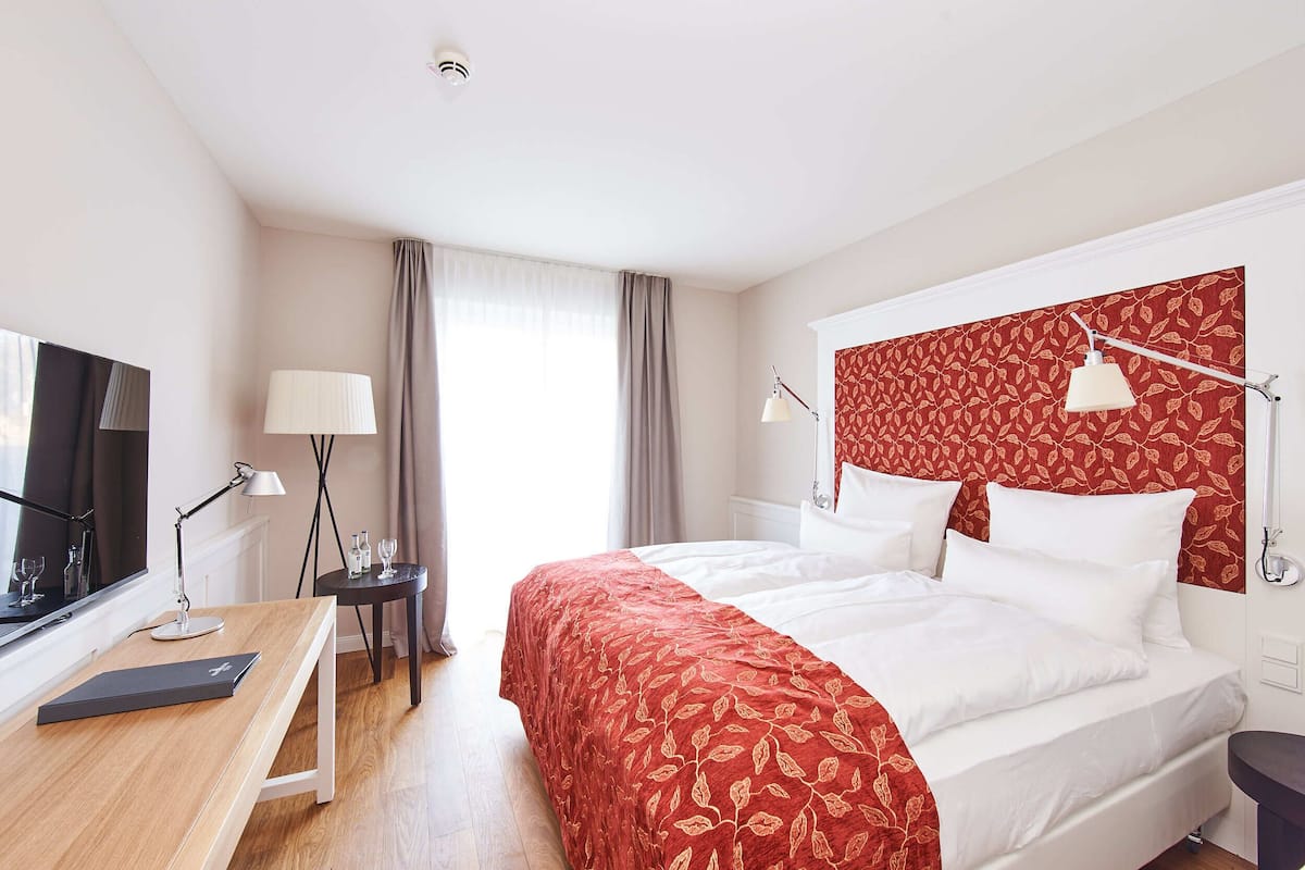 A hotel room with a bed, a desk, a TV, and a bedside table with a lamp.