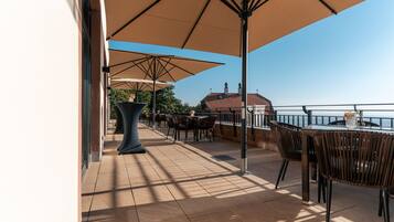 Une terrasse extérieure avec des tables et des chaises, de grands parasols et une vue sur un bâtiment situé de l’autre côté de l’eau.