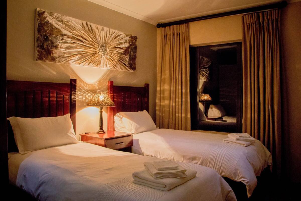 A hotel room with two beds, a nightstand with a lamp, a large abstract painting above the beds, and a view of the outside through a window with curtains.