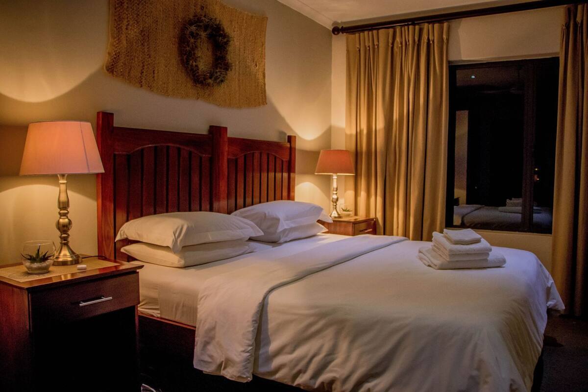 A hotel room with a wooden headboard, two bedside tables with lamps, a bed with white linens, a window with curtains, and a wall decoration.