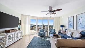 A living room with a large flat-screen TV, a ceiling fan, a sofa, a coffee table, and a view of the outdoors through large glass doors. - Recently Renovated Two Bedroom With Gorgeous Water Views! Complimentary Boat Slip! (Orange Beach)