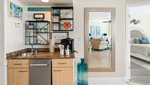 A modern kitchen with granite countertops, a dishwasher, and a coffee maker. There are shelves with glassware and decorative items. A large mirror reflects the living room with a sofa and a bed. - Recently Renovated Two Bedroom With Gorgeous Water Views! Complimentary Boat Slip! (Orange Beach)