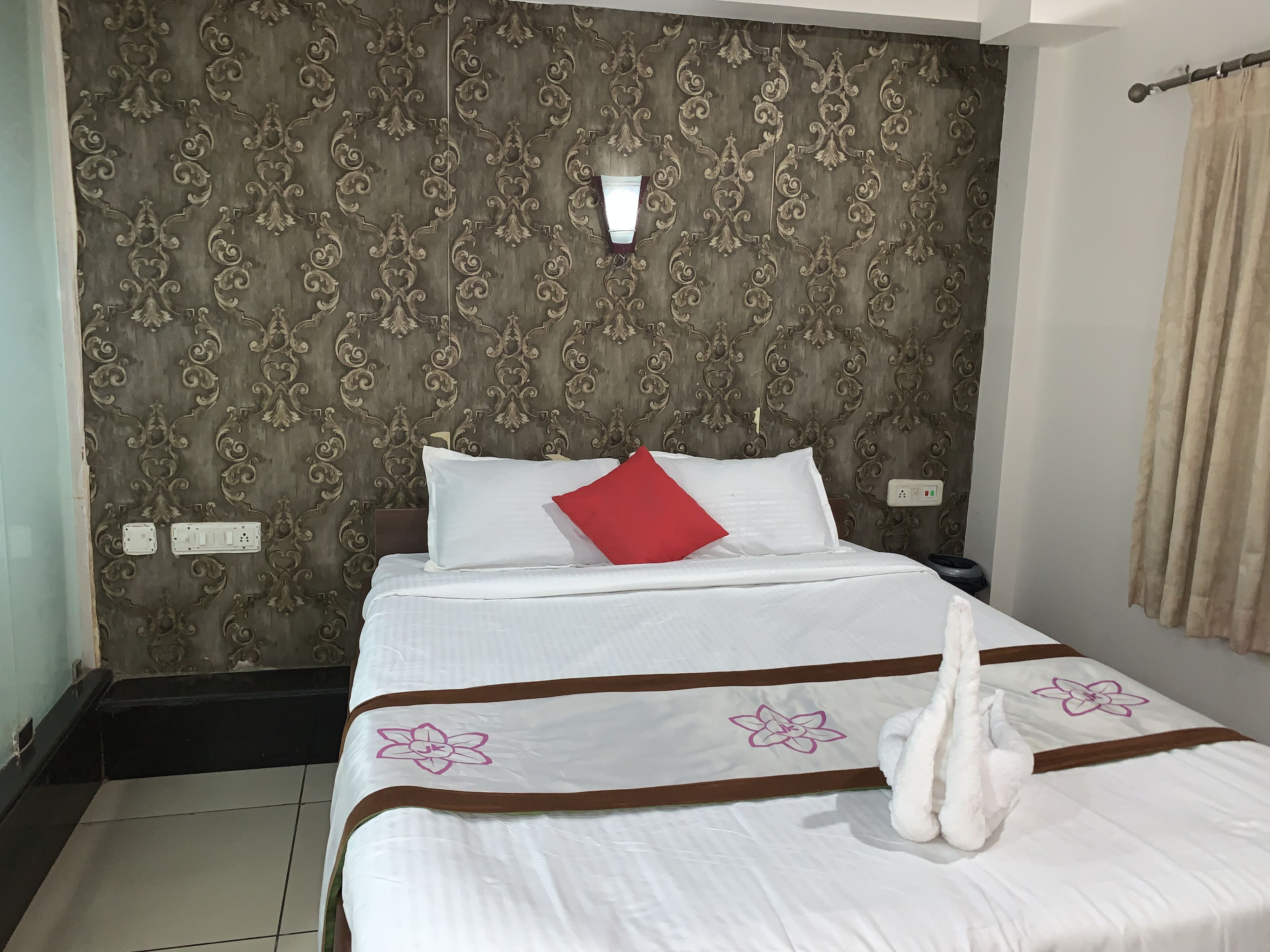 A neatly made bed with white bedding and a red pillow, a wall with ornate patterned wallpaper, and a light switch on the wall.