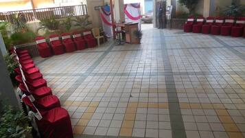 A covered outdoor seating area with red chairs and a small table, surrounded by tiled flooring and plants.