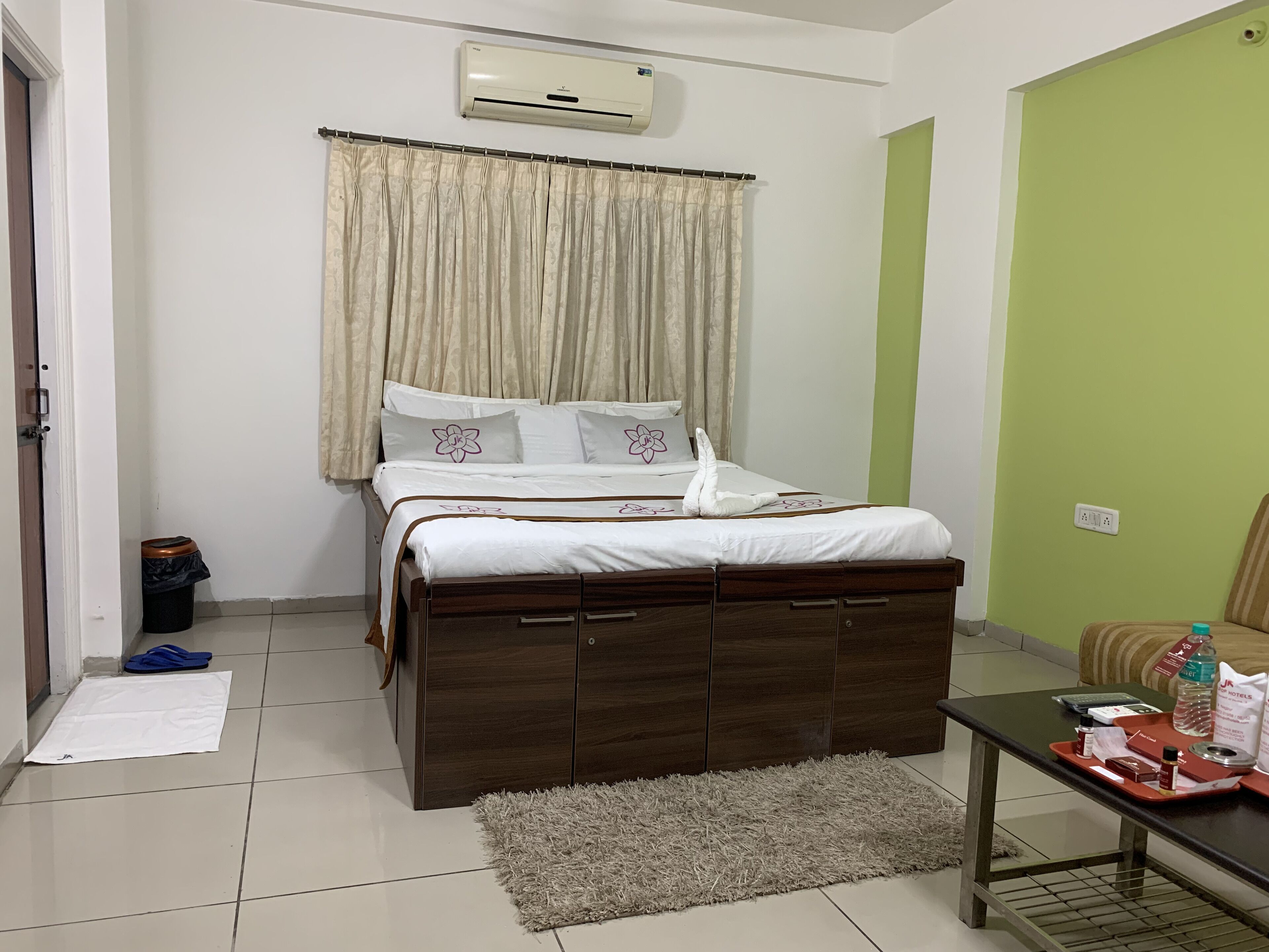A neatly arranged bedroom with a bed, a bedside table with toiletries, a trash bin, and a green wall.