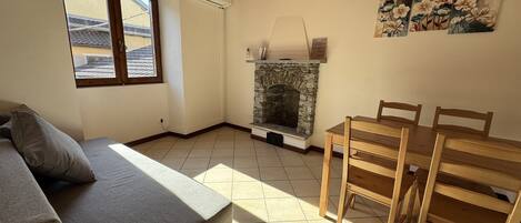 A room with a stone fireplace, a wooden dining table, a grey sofa, and a window with a view of buildings.