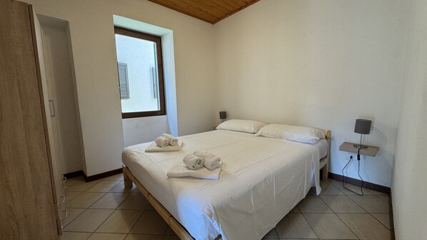 A bedroom with a bed, two pillows, a bedside table with a lamp, and a window.