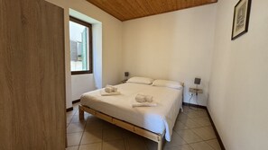 A bedroom with a wooden bed frame, white linens, a bedside lamp, and a window with shutters.
