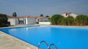 A rectangular swimming pool with a clear blue water surface, surrounded by a concrete deck. There are metal handrails along the edge of the pool. In the background, there are buildings with red-tiled roofs and white walls, and some greenery. - "La Fautaise" house in residence with swimming pool, near beach. (La Faute-sur-Mer)